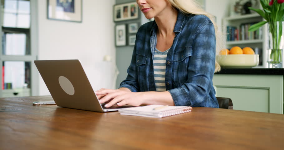 Tilt up shot of beautiful blond woman typing on her laptop and picking up her smart phone to send a text message. She is doing online browsing, checking her social media feed. Shot on RED Camera.