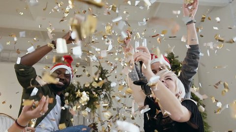 Happy young people in festive clothes with glasses of wine are smiling and dancing in flying gold confetti on New Year or Christmas party. Multi racial group of funny people celebrating party. Slow