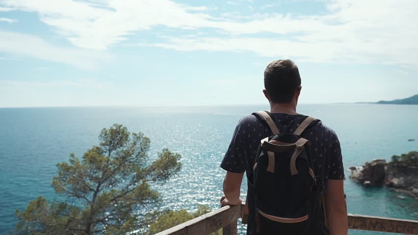 Shot from behind of a young male tourist enjoying beautiful view of a the sea, cliffs and forest. Guy with backpack on a walk.