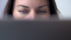 Close-up, caucasian girl with blue eyes works behind a laptop, half of the face is closed by the screen. Freelancer, hacker or user of social networks concept video.