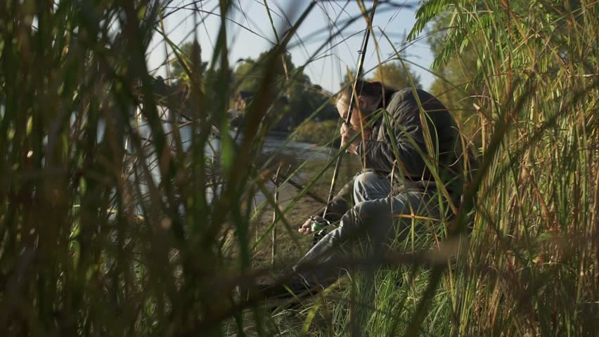 Fisherman is fixing his spinning sitting on river bank. The man is engaged in a fishing.