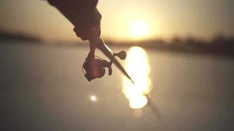 Male hand holds fishing rod on the background of the river during sunset close-up. Fisherman holds a fishing rod against the sunset. Sunlight through a fishing rod. River fishing.