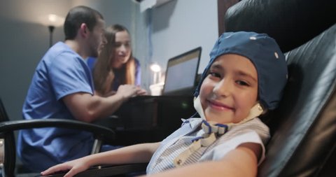 Portrait of young preteen autistic girl looking at camera in slow motion, while medical staff and mother reads EEG results on computer. The girl smiles while doing electroencephalogram