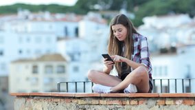 Surprised teenage girl finding online content on a smart phone sitting on a ledge on vacation