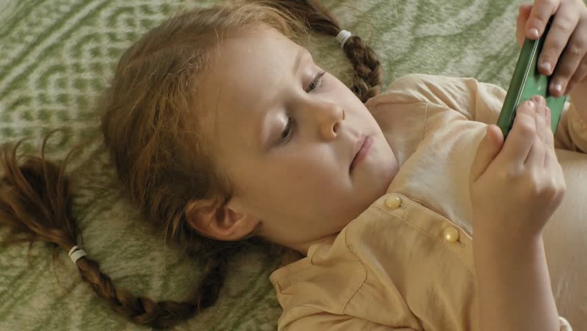 happy girl child with blond hair and pigtails, lying on the sofa, using the phone, smiling, portrait 4k