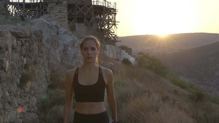 Young pretty light haired tanned girl with ponytail, slim body, flat stomach in short sport top and skinny leggings and sneakers walking fast on the mountains near fortress at sunset in slow motion