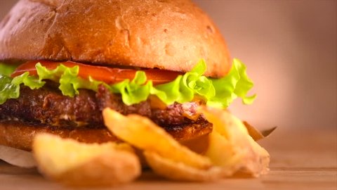 Hamburger with fries on wooden table closeup. Cheeseburger on fresh buns with succulent beef and fresh salad ingredients served with French Fries in on a rustic wooden table. 4K UHD slow motion video