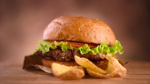Hamburger with fries on wooden table. Cheeseburger on fresh buns with succulent beef patties and fresh salad ingredients served with French Fries in brown paper on a rustic wooden table. 4K UHD