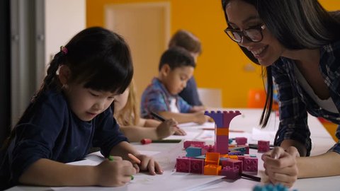 Lovely mixed race asian female teacher looking at drawing of little cute asian girl while helping class of preschool multi ethnic diverse children to draw on creative art lesson in kindergarten.