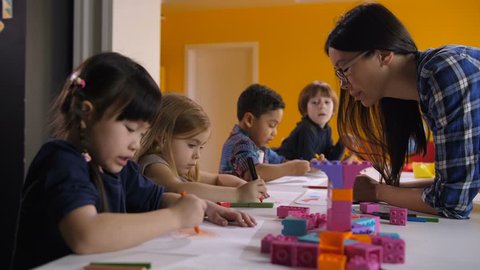 Cute diverse multi ethnic kids with asian teacher drawing at preschool class. Positive mixed race female teacher helping multiracial chilrdren with drawing during art lesson in kindergarten.