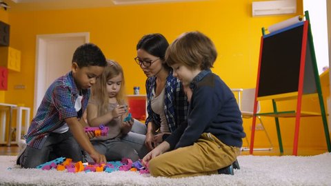 Joyful positive multicultural diverse children with attractive mixed race female teacher playing with colorful plastic building toy blocks in kindergarten, while sitting on floor in preschool class