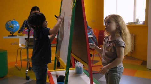 Creative cute multi ethnic girls drawing and writing with chalk and felt-tip pen on easel board in kindergarten while teacher with kids studying on background. Diverse kids learning at preschool class