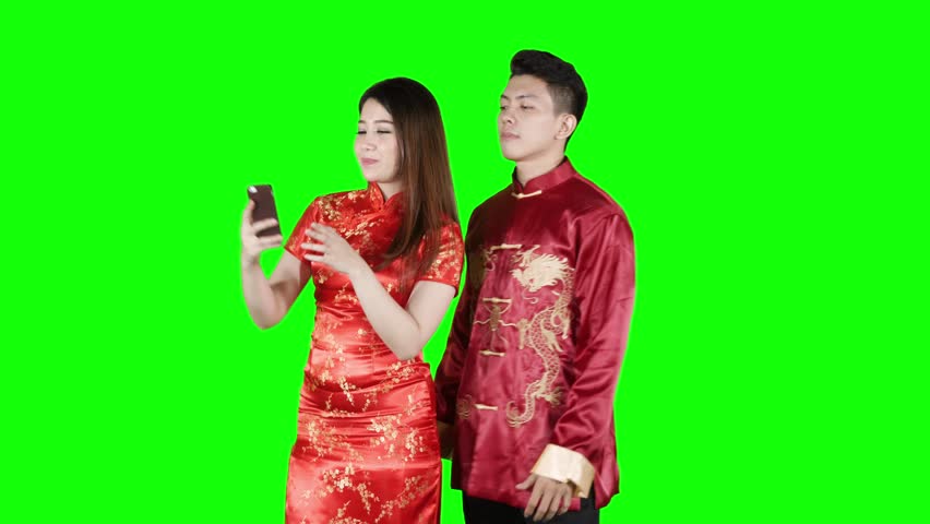 Happy Asian couple making video call with a mobile phone while wearing red cheongsam clothes in the studio. Shot in 4k resolution with green screen background