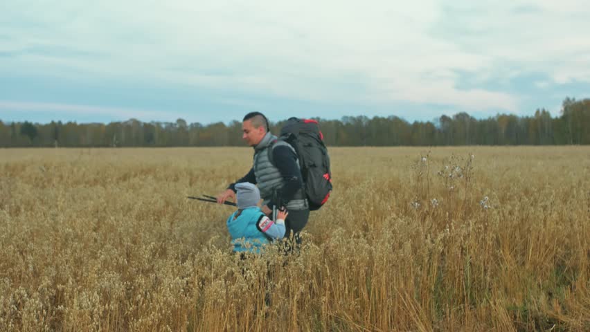 People walk near beautiful mountains in wheat field. Family travels. People environment by mountains, fields. Parents and kids walk using trekking poles. Man and woman have hiking backpacks, flasks