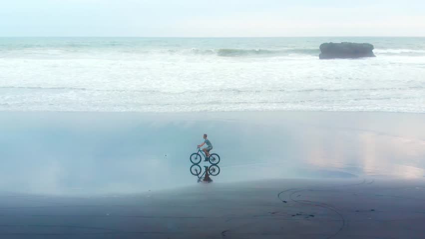 sporty young man riding his bike on the ocean beach on the background of big waves and sunset. Conceptual image of freedom, health and vivacity of spirit. Bali, Indonesia