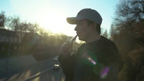 A young guy in a cap smokes an electronic cigarette at sunset, smoke burns in the sun, slow motion video