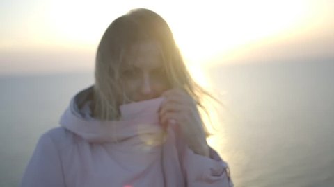 Young beautiful light haired girl posing sea background, wind blowing through her hair, she touching hair, posing, smiling tenderly, looking enigmatically at the camera, hiding nose in coat's collar, 