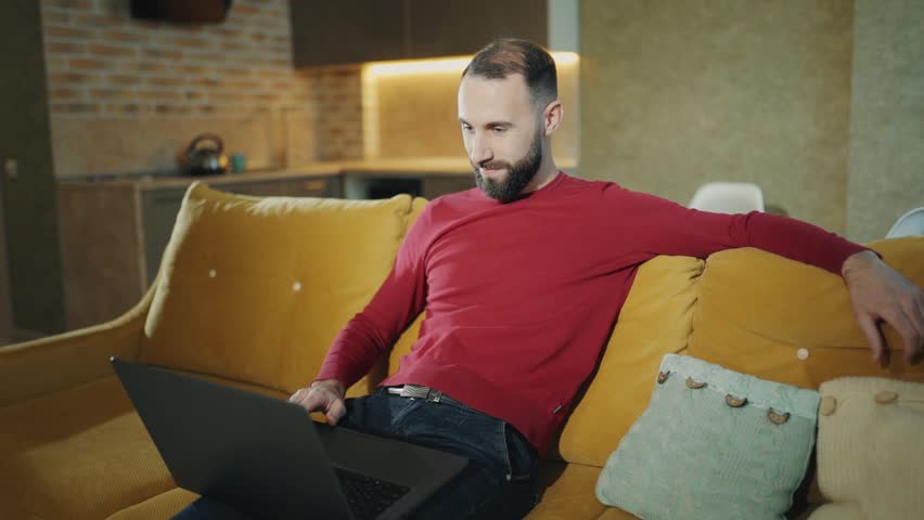 Happy bearded man is working at home with laptop and making a successful deal or win big money. Money falls from above and he is rejoices at home. Freelancer or businessman Royalty-Free Stock Footage #1021081243