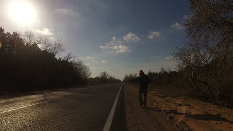 Mature man, tourist hitchhiker walks along road with a backpack on his shoulders, lifting his thumb up. Traveler is trying to stop car. Highway in summer forest