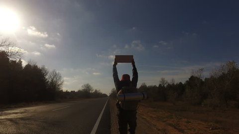 Mature man, tourist hitchhiker walks along road with a backpack on his shoulders, lifting tablet up. Traveler is trying to stop car. Highway in summer forest