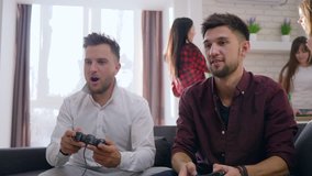 young gamers friends male play video games sitting on couch and funny girlfriends in background at home party