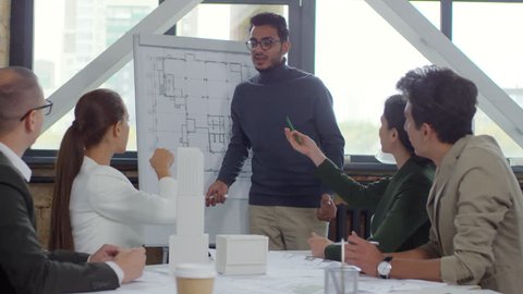Young middle eastern architect in eyeglasses explaining construction plan on flipchart to Caucasian colleagues while giving presentation during office meeting
