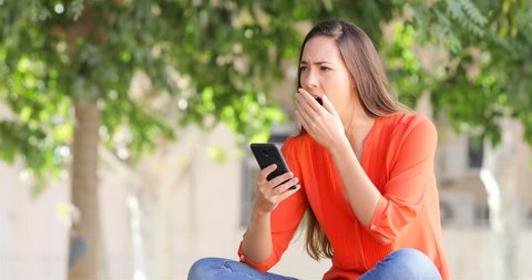 Woman yawning using a smart phone sitting on a bench in a park
