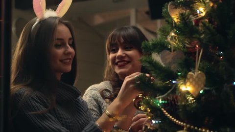 Girlfriends decorate christmas tree with joy and in festive mood. Two girls Laughing while dressing up christmas tree. Sisters together fill the Christmas tree with toys, preparing for holidays