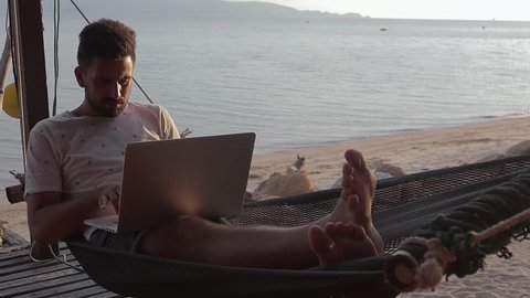Cinematic footage. A handsome young man working on his laptop in the hammock on the beach.