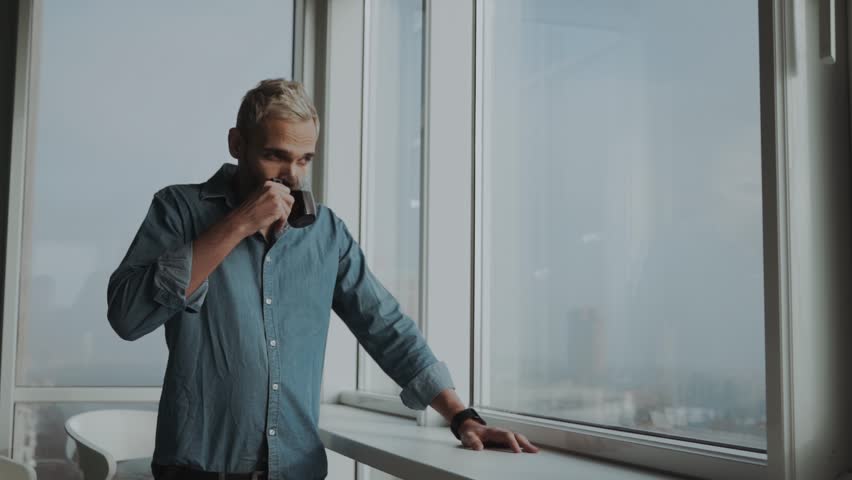 stylish man with a beard drinking tea / coffee and enjoying the view from the panoramic windows, modern office, panorama of the city Royalty-Free Stock Footage #1021106005