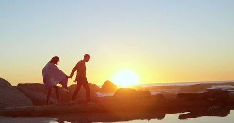 Couple walking with hand in hand on rocky shore. Sun setting in sea in the background 4k