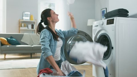 Beautiful and Happy Brunette Young Woman is Sitting Next to the Washing Machine in Homely Jeans Clothes. She Laughts and Throws Laundry Clothes into the Air. Living Room with Modern Interior.