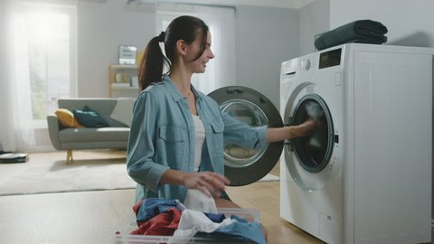 Beautiful Smiling Brunette Young Woman Sits in Front of a Washing Machine in Homely Jeans Clothes. She Loads the Washer with Dirty Laundry. Bright and Spacious Living Room with Modern Interior.