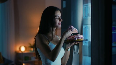 Beautiful Young Woman Comes to the Kitchen in the Evening. She is Hungry and Opens the Fridge. Takes out a Piece of a Delicious Creamy Cake and Bites It. She Feels Satisfied.