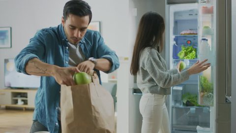Beautiful Young Couple Come to the Kitchen with Fresh Groceries in Brown Paper Bag. Man is Handing Fresh Salad Greens, Apples and Oranges to the Girl Who Puts Them in the Fridge.