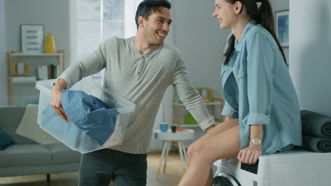 Beautiful Young Couple Talking in a Living Room at Home. Girl is Sitting on a Washing Machine, Man Holding Laundry. They are Happy and Laugh. Room has Modern Interior.