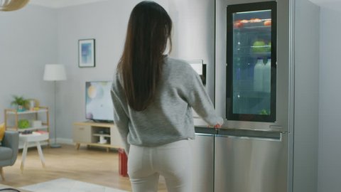 Beautiful Brunette Girl in White Jeans and Grey Sweater Comes to the High-Tech Fridge with a Glass Door and Takes a Green Apple. She goes to the Living Room. Flat has Modern Interior.