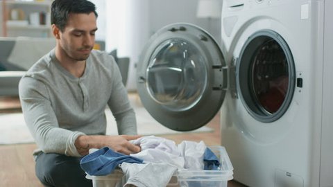 Handsome Smiling Young Man in Grey Jeans and Shirt Sits in Front of a Washing Machine at Home. He Loads the Washer with Dirty Laundry. Bright and Spacious Living Room with Modern Interior.