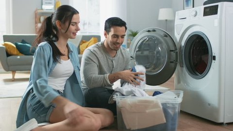 Beautiful Young Couple Sit Next to a Washing Machine at Home. They Laugh and Have a Friendly Argument While Loading the Washer with Dirty Laundry. Bright and Spacious Living Room with Modern Interior.