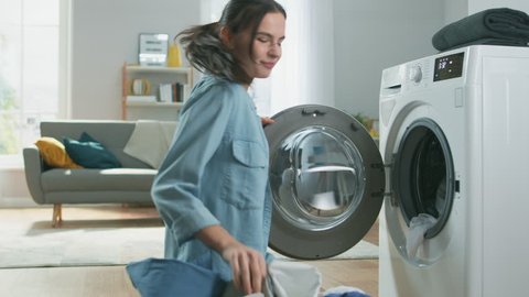 Beautiful and Happy Brunette Young Woman Dancing and Moving Towards the Washing Machine in Homely Jeans Clothes. She Loads the Washer with Dirty Laundry. Spacious Living Room with Modern Interior.