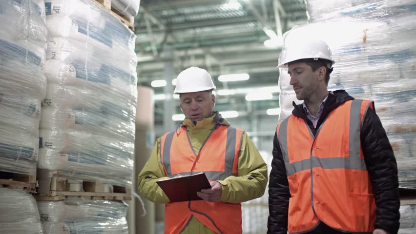 Workers walking and discussing stock inventory against tall shelves, in a warehouse with bright lamps. Royalty-Free Stock Footage #1021118842