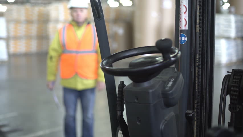 Concentrated adult forklift driver in white hardhat and orange workwear coming inside, checking the tablet and making notes inside machine. Royalty-Free Stock Footage #1021118845