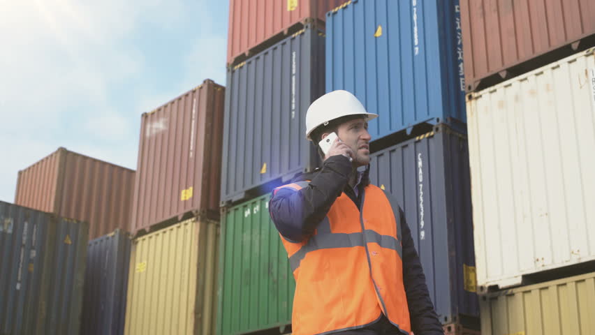 Caucasian man in white hardhat talking by cellphone standing in dockyard containers. Royalty-Free Stock Footage #1021118911