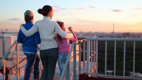 Back of mother and two children stand on roof of tall building and look at sunset