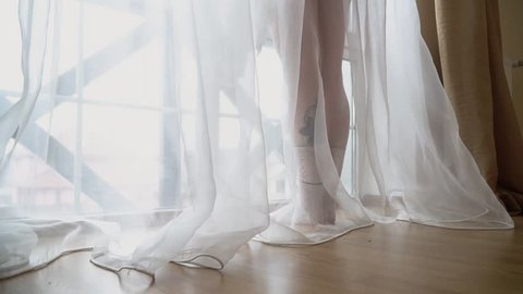 A woman throws her nightie on the floor standing near a large bright window. Naked woman in the bedroom.