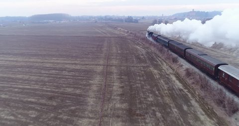 Old train: aerial view of steam train running on the tracks in the countryside. Heritage historic steam locomotive with white smoke seen from above by drone.
