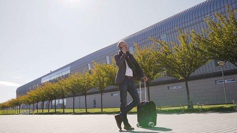 Young stylish man talking on his phone standing with suitcase while exiting the airport. Outdoors. Business style, traveler, modern lifestyle, business trip. Slow motion