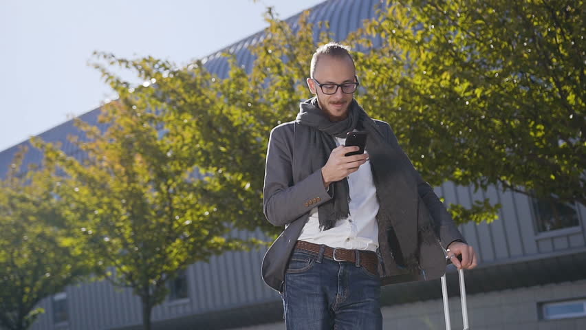 Young bearded businessman dressed in jacket is talking on phone while he standing near the modern office building. Outdoors . Concept: office worker, business, business trip, advertising. Slow motion