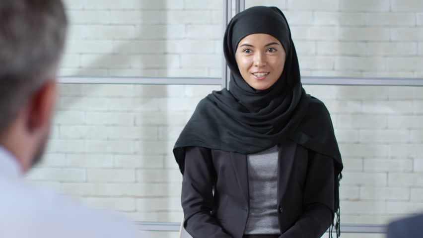 Tracking shot of cheerful Muslim woman in hijab sitting opposite businessman with beard and elderly manager with grey hair and talking about her professional skills during panel job interview