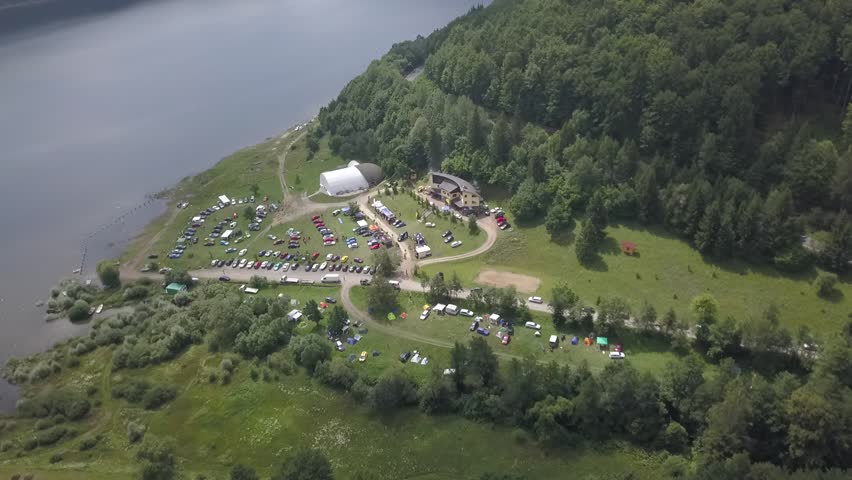 Drone footage of a car meeting near a big lake, surrounded by forest